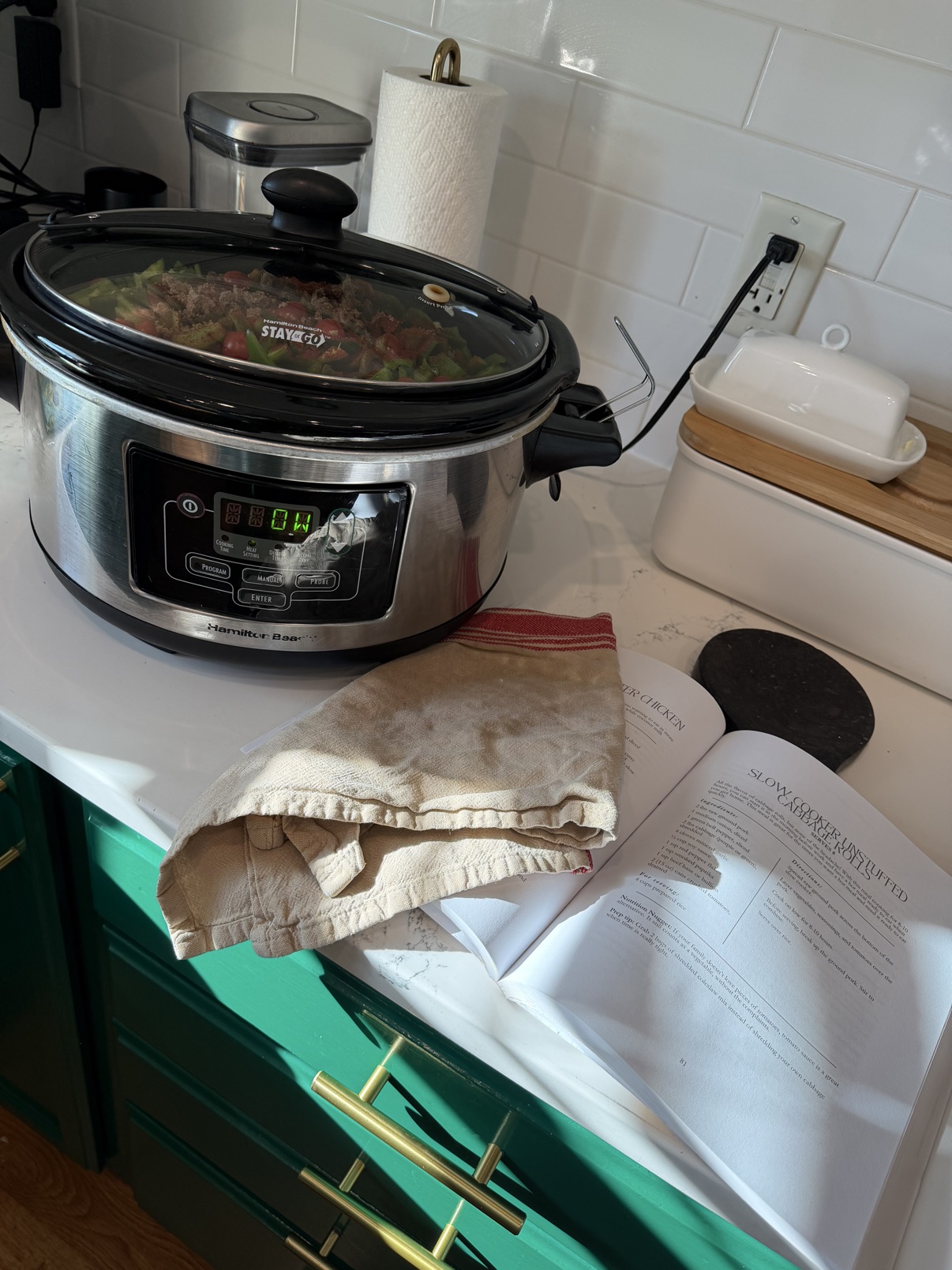 Slow cooker filled with unstuffed cabbage rolls ingredients beside the Uncomplicate Your Plate cookbook open to page 31