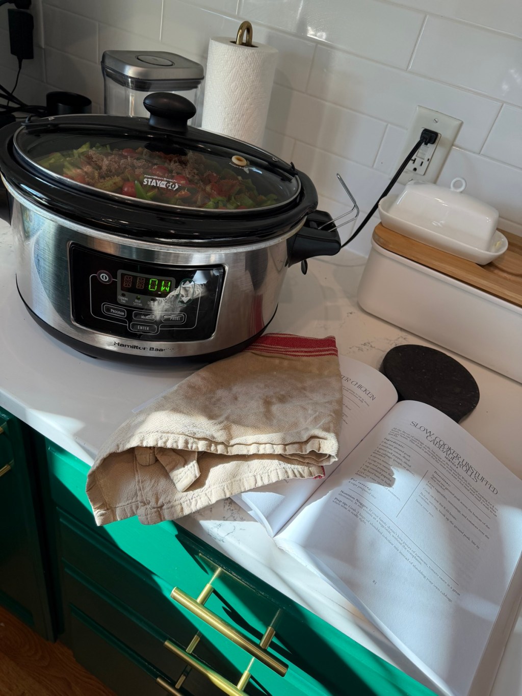 Slow cooker filled with unstuffed cabbage rolls ingredients beside the Uncomplicate Your Plate cookbook open to page 31